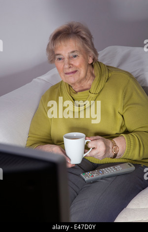 Senior lady da solo guardando la tv tenendo una tazza, il telecomando sul suo grembo. Foto Stock