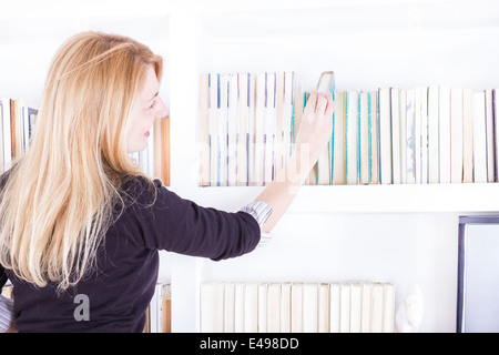 Donna bionda tirando un libro della biblioteca off ripiano, ragazza selezionando Rubrica da scaffale Foto Stock