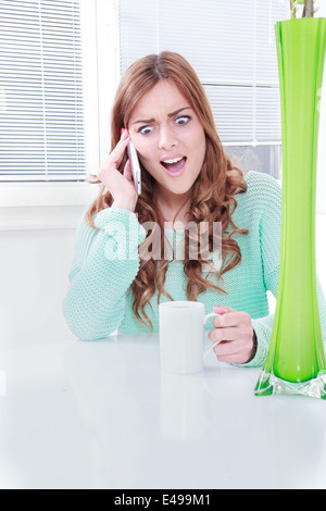 Giovane donna sul telefono cellulare con reazione negativa, ragazza seduta a tavola con la tazza di caffè parlando al telefono cellulare audizione cattive notizie, scioccato donna con espressione faccia tenendo il telefono cellulare Foto Stock