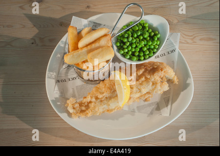 Pesce e patatine (Eglefino nella pastella) con piselli e un segmento di limone su una piastra bianca su di un tavolo di legno. Foto Stock