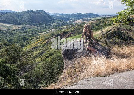 Donna bionda che indossa abiti di oziare sulla roccia guardando fuori in campagna valle guardando in lontananza Foto Stock