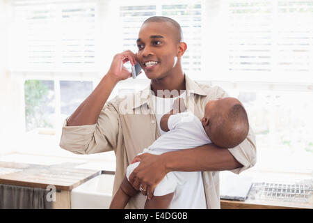 Padre Felice alimentando il suo bambino una bottiglia mentre si è al telefono Foto Stock