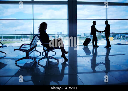 Profilo di imprenditrice seduto in aeroporto su sfondo dei suoi colleghi lo handshaking mediante la finestra Foto Stock