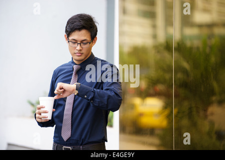 Ritratto di imprenditore cinese in piedi vicino edificio per uffici a Panama con tazza di caffè e controllando il tempo di guardia Foto Stock