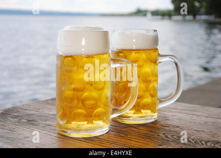 Due boccali da birra con birra fresca e schiuma su una tavola di legno in un giardino della birra sul lago, vicino fino Foto Stock