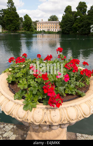 Rosso dei gerani crescente nell urna vaso contenitore con vista verso il Museo non 1 oltre la Casa delle Palme Pond. I Giardini di Kew. Kew, West London REGNO UNITO. Foto Stock