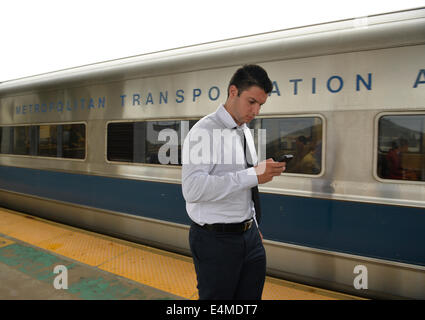 Merrick, New York, Stati Uniti - 14 Luglio 2014 - in serata durante le ore di punta, un giovane uomo nel business vestiti lascia il treno e guarda al suo smart phone mobile device come egli cammina sulla piattaforma sopraelevata di Merrick stazione ferroviaria di Babilonia il ramo, dopo MTA Metropolitan Transit Authority e Long Island Rail Road unione parla deadlock, con potenziale sciopero LIRR aleggiano pochi giorni prima. Credito: Ann e Parry/Alamy Live News Foto Stock