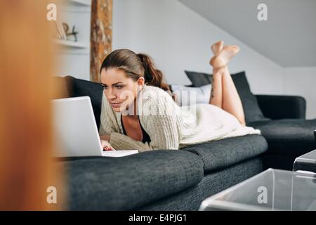 Immagine della giovane donna occupato con un portatile a casa. Caucasian modello femminile sdraiato sul lettino lavorando sul computer portatile. Foto Stock