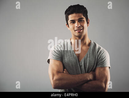 Ritratto di bel giovane uomo in piedi con le braccia incrociate contro uno sfondo grigio con copia spazio. Ispanico moda maschile mod Foto Stock