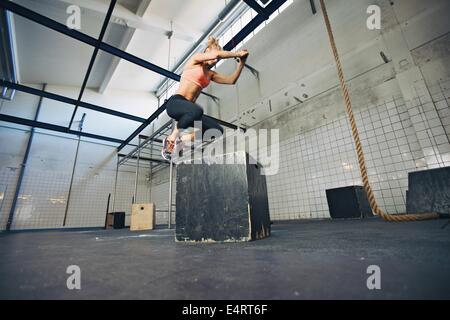 Basso angolo vista del giovane atleta femminile box jumping in una palestra crossfit. Montare la donna sta eseguendo la casella salta in palestra. Foto Stock