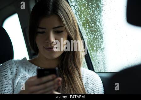 Giovane donna seduta all'interno auto e guardando il telefono cellulare sul giorno nuvoloso, Germania Foto Stock