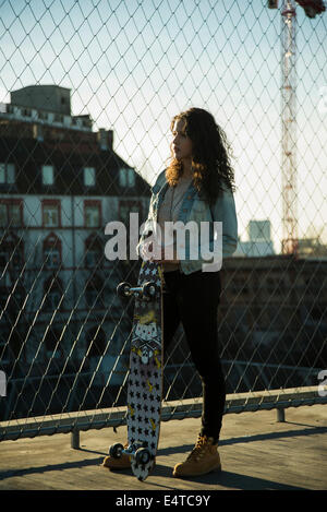 Ragazza adolescente in piedi all'aperto accanto alla catena collegamento recinto vicino comercial dock, tenendo sullo skateboard, Germania Foto Stock