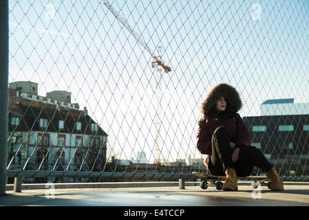 Ragazza adolescente indossando cappotto invernale, seduti su skateboard all'aperto, accanto alla catena collegamento recinto vicino comercial dock, Germania Foto Stock