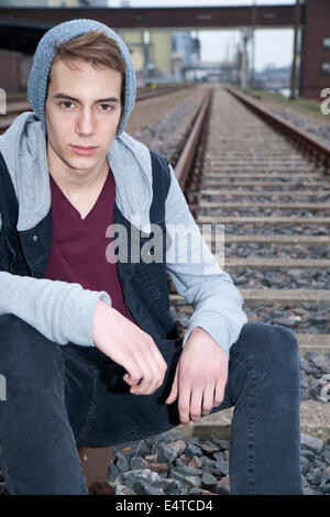 Ritratto di ragazzo adolescente seduto sui binari della ferrovia, guardando la telecamera, Germania Foto Stock