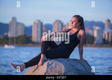 Un attraente giovane donna si rilassa su una roccia con l'oceano e gli edifici di Vancouver dietro di lei. Foto Stock