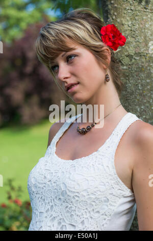 Attraente giovane donna in un elegante bianco lacy top con red rose nei suoi capelli come lei si appoggia contro un albero nel giardino Foto Stock