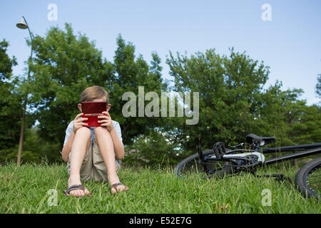 Un giovane ragazzo seduto sull'erba a giocare una mano di gioco elettronico. Foto Stock