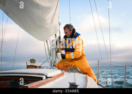 Sailor in giallo le intemperie marcia tirando la drizza per sollevare la randa come egli si dirige verso le nuvole nella sua barca a vela Foto Stock