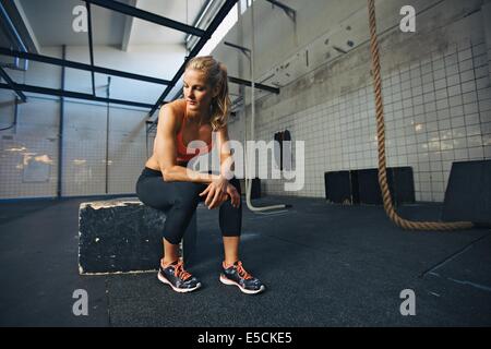 Caucasian atleta donna seduta su una casella nella palestra di pensare. Montare la giovane donna prendendo pausa dopo il suo allenamento in palestra. Foto Stock