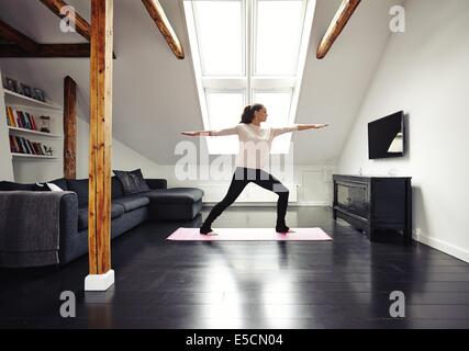 Lunghezza completa immagine di montare la donna in piedi sul tappeto di esercizio con le braccia aperte facendo yoga in camera amorevole. Caucasian modello femminile Foto Stock