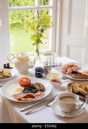 Prima colazione inglese tradizionale tabella di cui fuori dalla finestra sala interna con tè toast pancetta, salsicce e uova sulla tovaglia Foto Stock