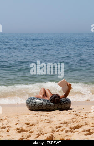 Donna che si rilassa in vacanza, leggendo un libro sulla spiaggia al bordo delle acque, Algarve, Portogallo Europa Foto Stock