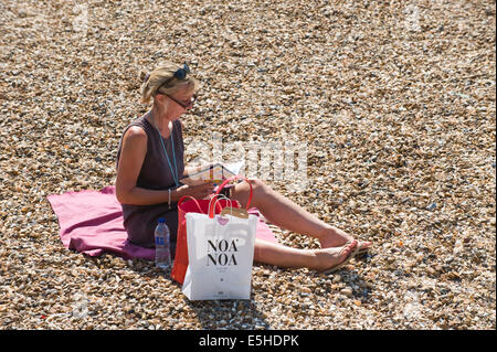 Donna di mezza età lettura Oyster Festival programma seduti al sole sulla spiaggia a Whitstable Kent England Regno Unito Foto Stock