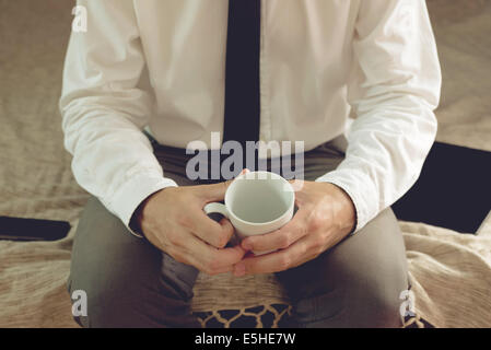 Imprenditore solitario nella camera di albergo di seduta sul letto e tenendo tazza vuota Foto Stock