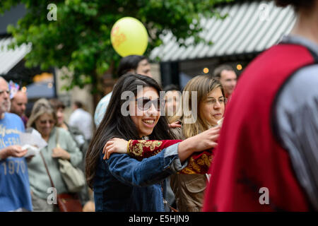 Giovane donna attraente in occhiali da sole si unisce con un greco ballo folk troupe, Marylebone street Fayre fiere estive, Londra, Regno Unito. Foto Stock