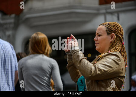 Una giovane donna con brunette capelli intrecciati prende una foto sul suo cellulare sulla strada a Marylebone Summer Fayre, Londra, Regno Unito. Foto Stock