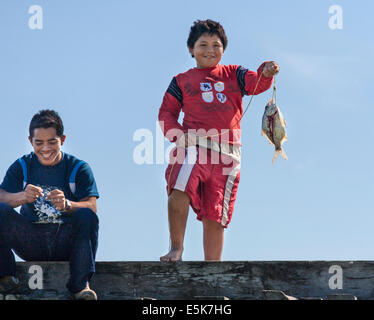 La visualizzazione delle catture. Un ragazzo visualizza un solo pesce pescato mentre un altro ragazzo ride. Entrambi sono sul bordo di un ponte di legno Foto Stock