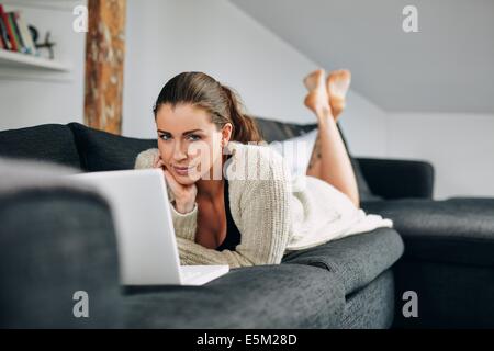 Ritratto di bella giovane femmina sdraiato sul divano con un laptop guardando la fotocamera. Rilassata giovane donna sul lettino con il computer portatile. Foto Stock