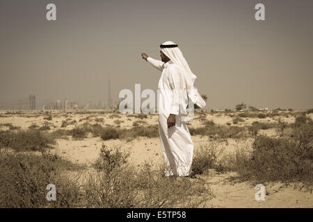 Uomo arabo in abito nazionale si erge nel deserto e guarda alla città di Dubai Foto Stock