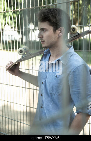 Giovane serio con lo skateboard in spalla dietro il recinto del parco Foto Stock