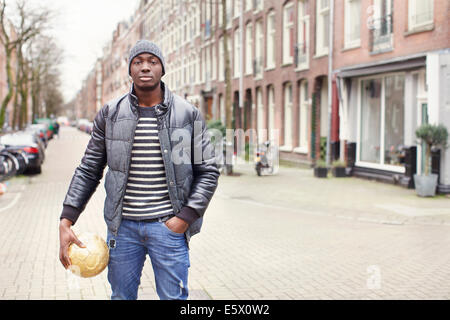 Ritratto di giovane uomo su strada tenendo palla calcio, Amsterdam, Paesi Bassi Foto Stock