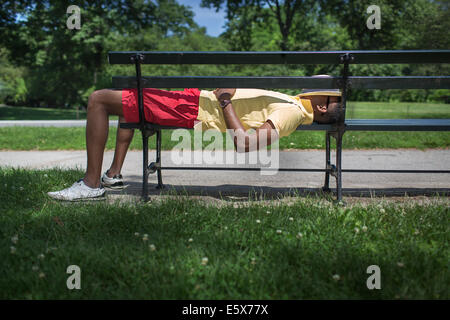 Vista laterale del giovane uomo disteso su una panchina nel parco con fedora che ricopre la faccia Foto Stock