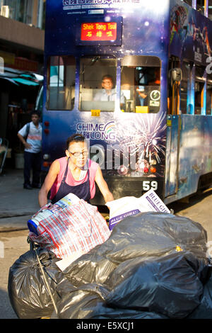 Femmina cinese Street Cleaner spinge un carrello pesante di rifiuti attraverso Chun Yeung Street, Vintage Hong Kong Tram in background. Foto Stock