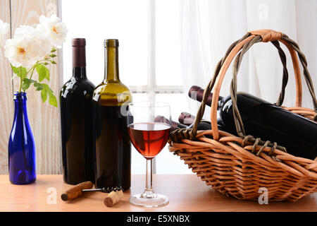 Vino ancora in vita con un paniere di bottiglie e un bicchiere di vino rosso e bianco rose in un vaso su un tavolo di fronte a una finestra con Foto Stock