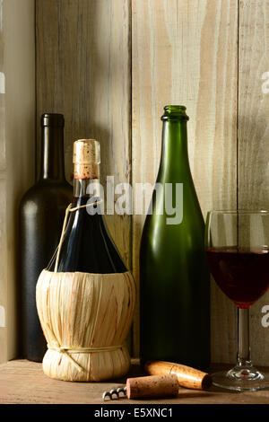 Un vino ancora in vita con una luce calda da una finestra sul lato. Tre bottiglie, un bicchiere di vino e vite di sughero in un ambiente rustico, Ver Foto Stock