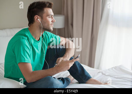 Giovane uomo seduto sul letto ascoltando la musica con lo smartphone Foto Stock