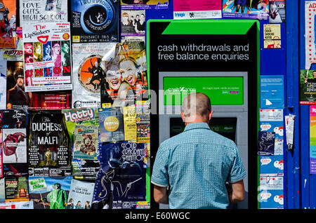 Un giovane uomo con un bancomat in Grassmarket circondato da manifesti con mostra sull'Edinburgh Festival Fringe. Foto Stock