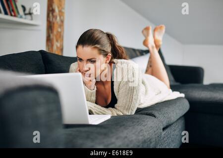 Ritratto di giovane e bella donna che lavorano su computer portatile mentre sdraiato sul divano. Utilizzando femmina portatile a casa. Foto Stock