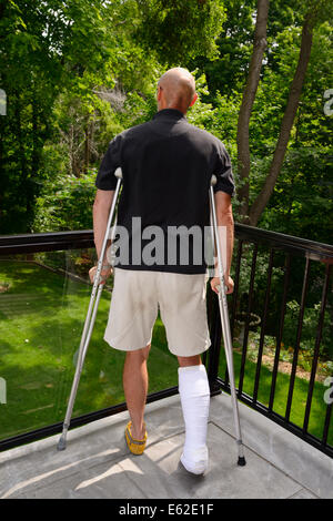 Uomo ferito ad una gamba in un cast e stampelle recupero da lesioni su un balcone che guarda a una foresta Foto Stock