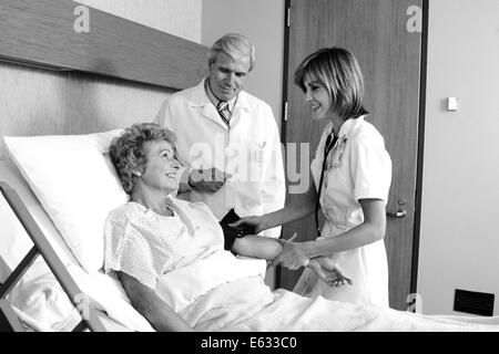 Anni ottanta donna anziana nel letto di ospedale avente la pressione del sangue preso dalla femmina maschio infermiere medico in piedi accanto a lei Foto Stock
