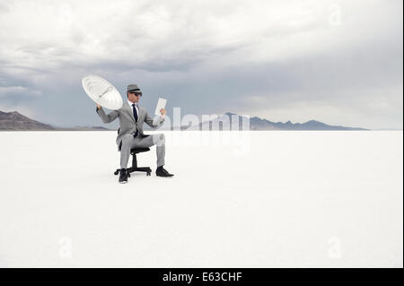 Imprenditore seduti all'aperto nel drammatico paesaggio deserto tenendo la comunicazione via satellite e ricevitore digitale compressa Foto Stock