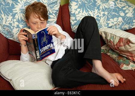 9 anni di vecchio ragazzo lettura Scorpia e romanzo di Anthony Horowitz sul divano a casa dopo la scuola Foto Stock