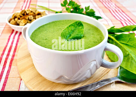 Zuppa verde purea in una ciotola con il verde di spinaci in foglia, cucchiaio sul bordo, crostini, prezzemolo su uno sfondo di stoffa Foto Stock