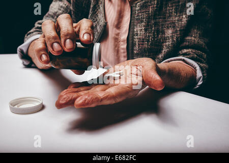 Tagliate il colpo di una vecchia donna seduta al tavolo agitando una pillola al di fuori di un vaso di pillola. Concentrarsi sulle mani. Femmina Senior tenendo medicin Foto Stock