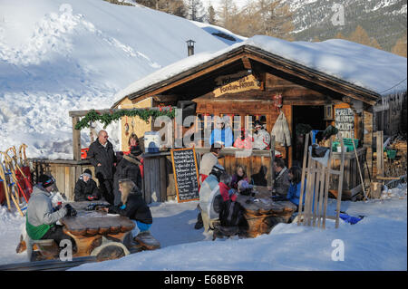 Piccolo ristorante con fonduta svizzera. I clienti stanno avendo un drink all'aperto nella neve approfittando degli ultimi raggi di sole Foto Stock