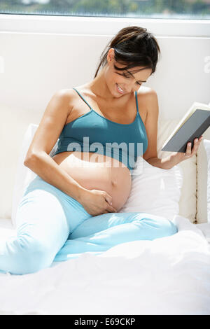 Incinta donna ispanica lettura a letto Foto Stock